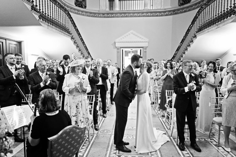 B&W image of bride and groom kissing with their friends and family clapping as they are announced husband and wife.