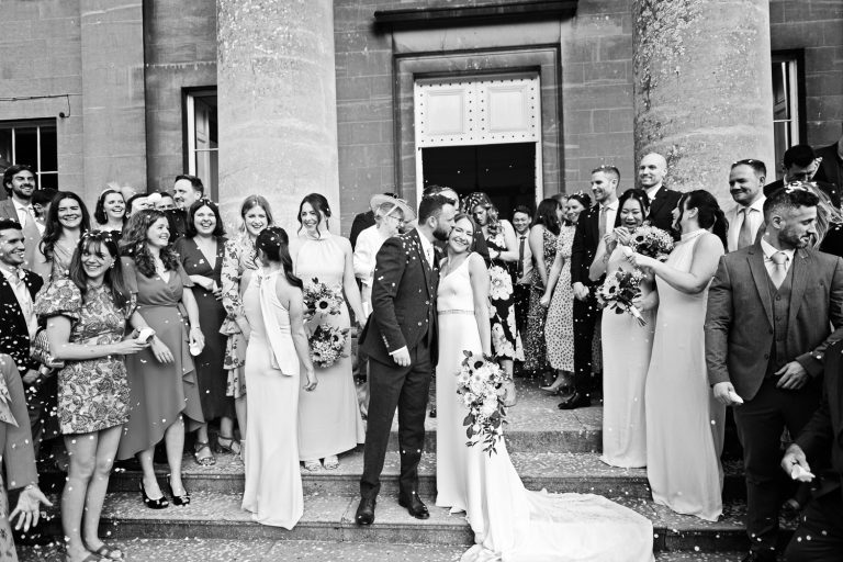 B&W classic photo of groom kissing the bride on the steps with their family and friends surrounding them after being showered with confetti.