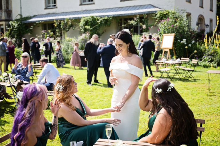 A bride chats to her bridesmaids during the wedding reception. Wedding guests behind them in the gardens in front of the Main House at the Matara.