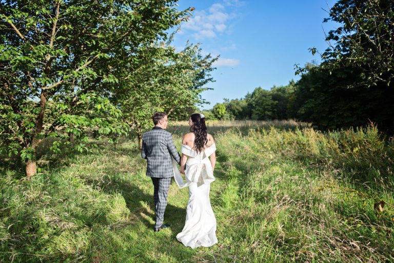 Bride and groom hold hand in hand through the long grass fields typical of an English countryside of yesteryear.