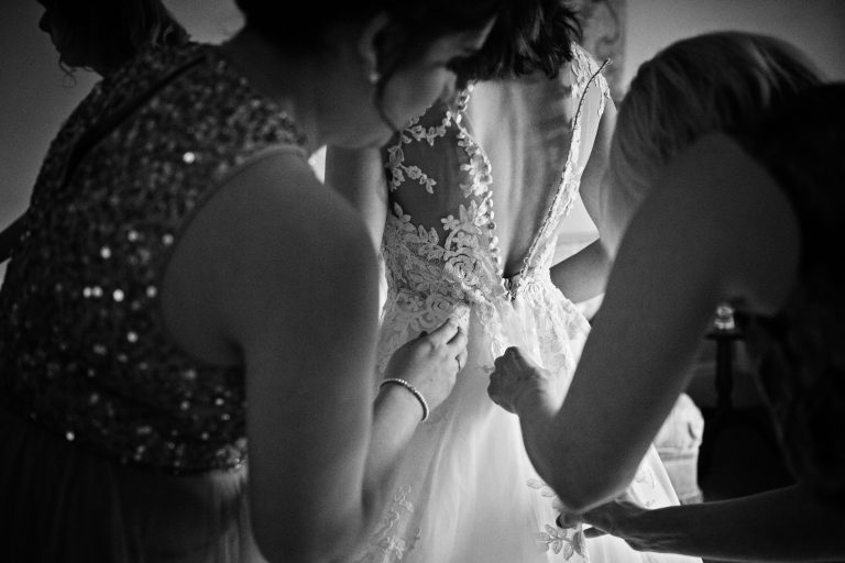 B&W photo of bride having her wedding dress done up by her bridesmaids.