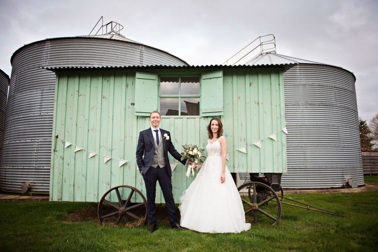 Bride and groom hold the wedding flowers and stand in front (smiling facing the camera) of a wooden pastel green cart. Large silo's behind them.
