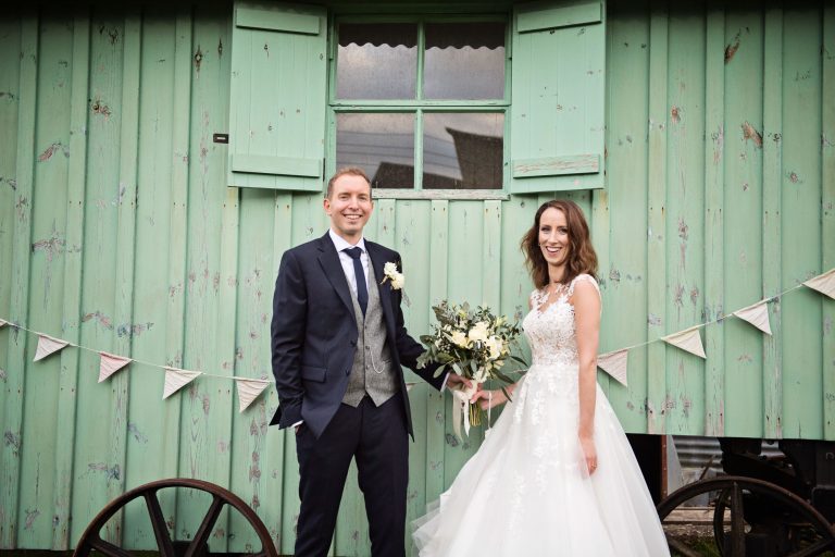 Bride and groom hold the wedding flowers and stand in front (smiling facing the camera) of a wooden pastel green cart.