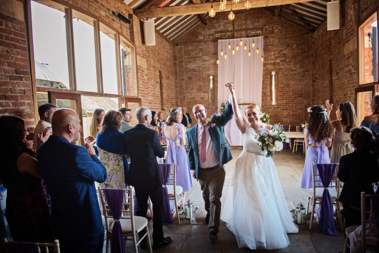 Bride and groom celebrating by their hands in the air as they walk down the wedding isle at Mickleton Hills Farm.