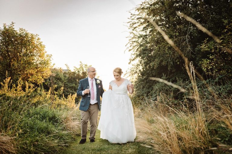 Bride and groom walk through the long grass at golden hour.