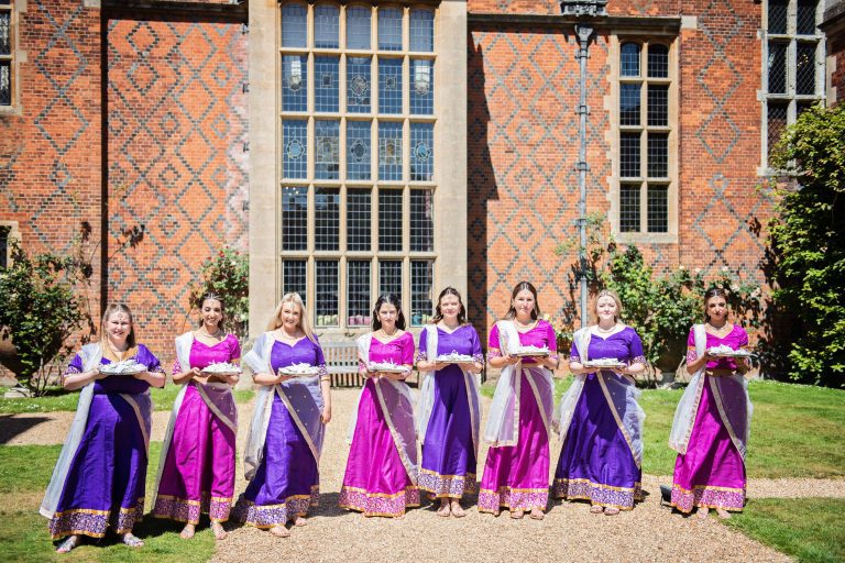 Bridesmaids wearing colourful sari's and holding wedding gifts stand in line.