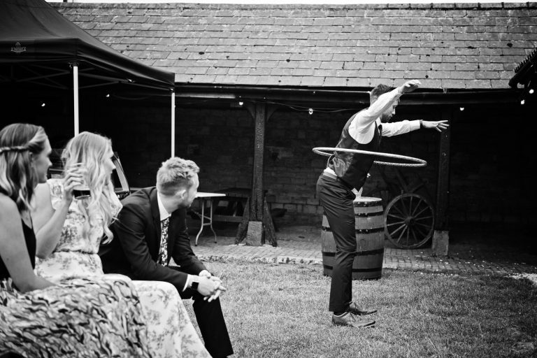 B&W image of guest doing the hula hoop in his wedding attire. Other guests watching him.