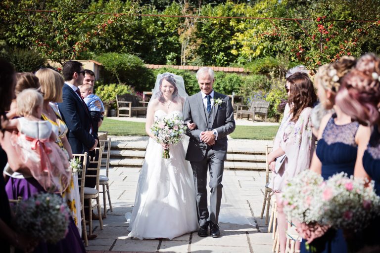 Bride walks down the wedding isle with her father. Wedding guests look onwards