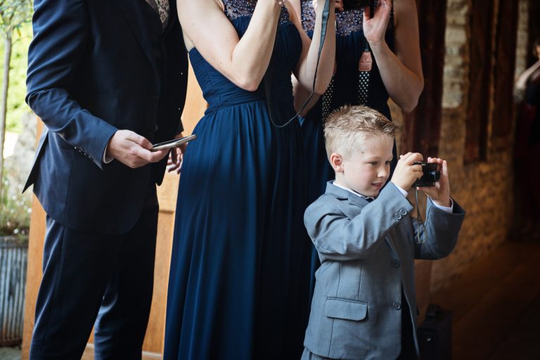 Little boy taking photo of something (bride and groom!), with wedding guests clapping behind him.