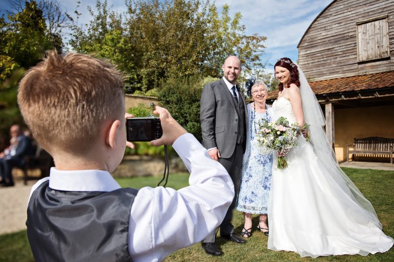 Little wedding guest taking a photograph of the bride and groom standing with the grooms mum.