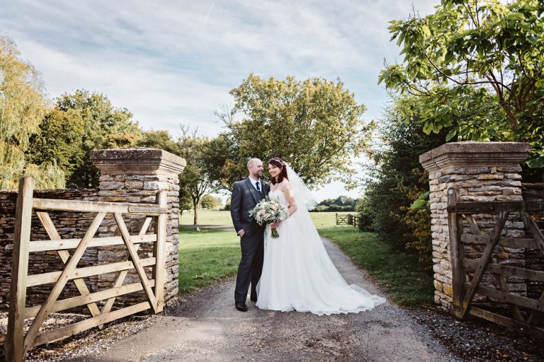 Relaxed photo of the bride and grooms standing in in between an opened gate that leads onto a driveway in a farmland setting.