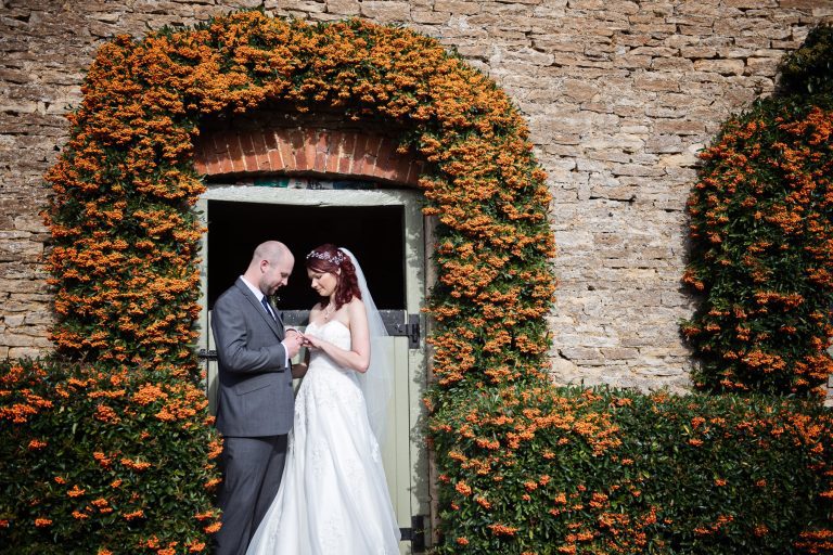 Bride and groom look at each others new wedding rings whilst they are stood in front of a farm door. A beautiful shaped orange pyracantha surrounds them.