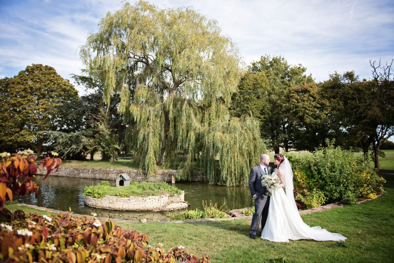 Bride and groom stand chatting to each other in front of a small lake and willow tree.