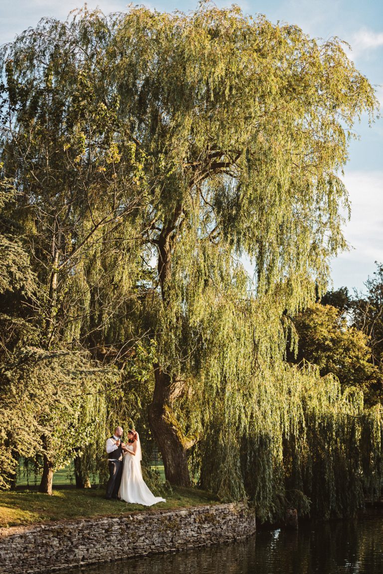Bride and groom chat wilst stood next to a large willow tree.