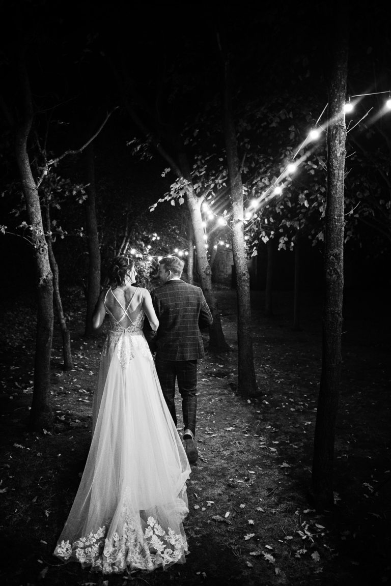 Portrait B&W photo of bride and groom walking down a festoon lit path. Atmospheric. Natural.