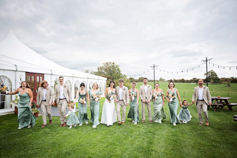 Bridal party walk hand in hand on the grass in front of the marquee.