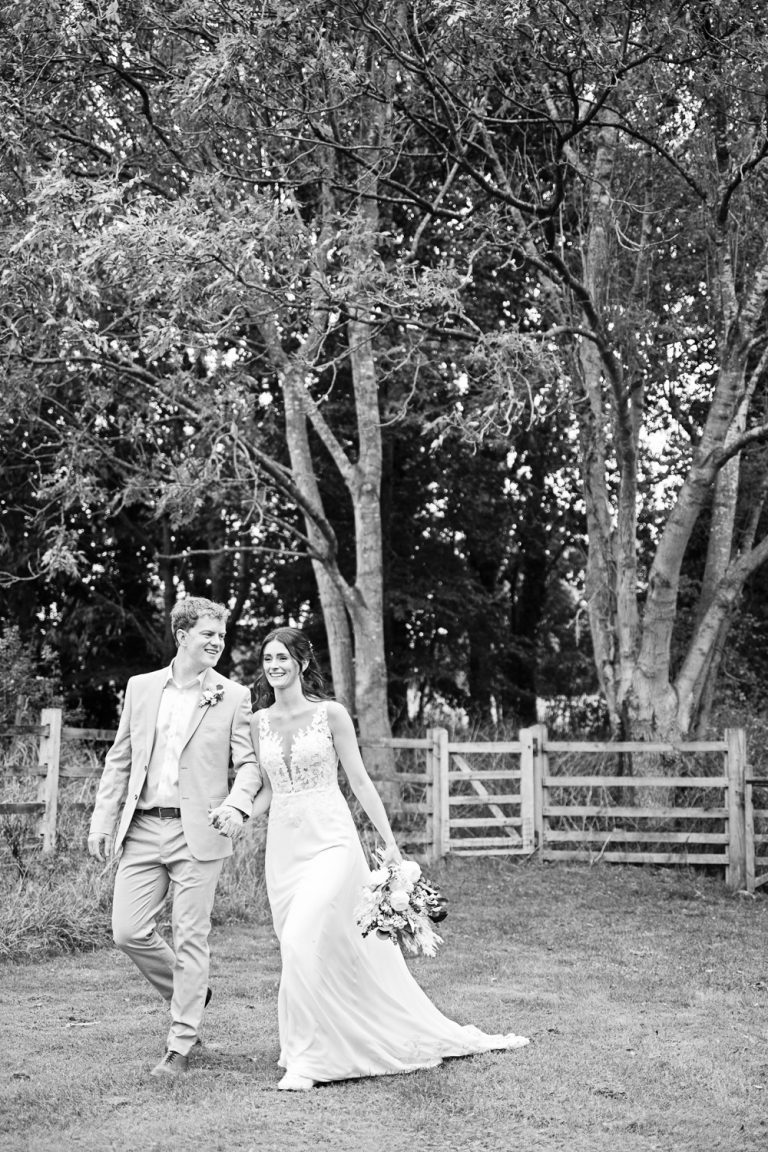 B&W portrait candid photograph of bride and groom walking hand in hand away from a gate and woodland.
