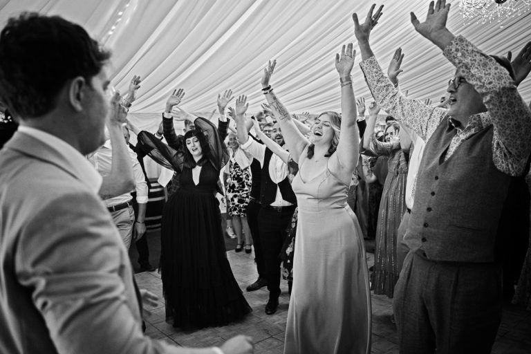 B&W fun candid photograph of lots wedding guests dancing with their hands in the air. Think Michael Jackson "Thriller".