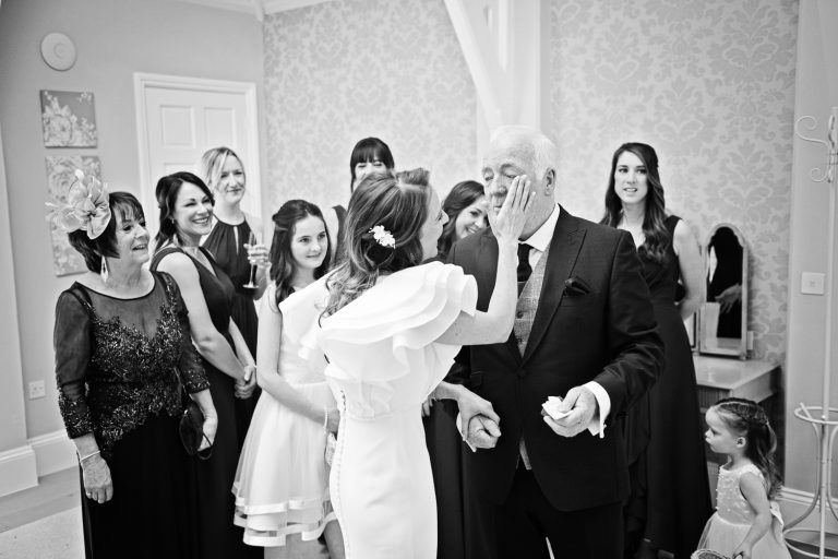 Candid storytelling photo of the bride loving her dad by wiping a tear away from his eye. Team bride watching behind.