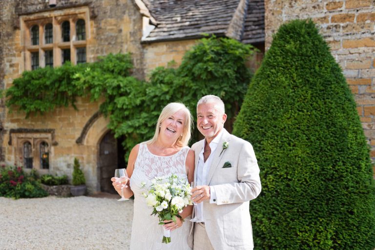 Bride and groom stand outside the front of Temple Guiting Manor with a glass of wine/fizz each. Looks like an Omaze photo!