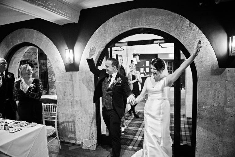 B&W photo of bride and groom entering the wedding breakfast room cheered on by friends and family.