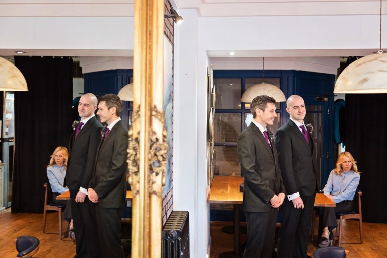 Groom and best man stand at the front of the wedding isle reflected by a mirror before his bride walks down.