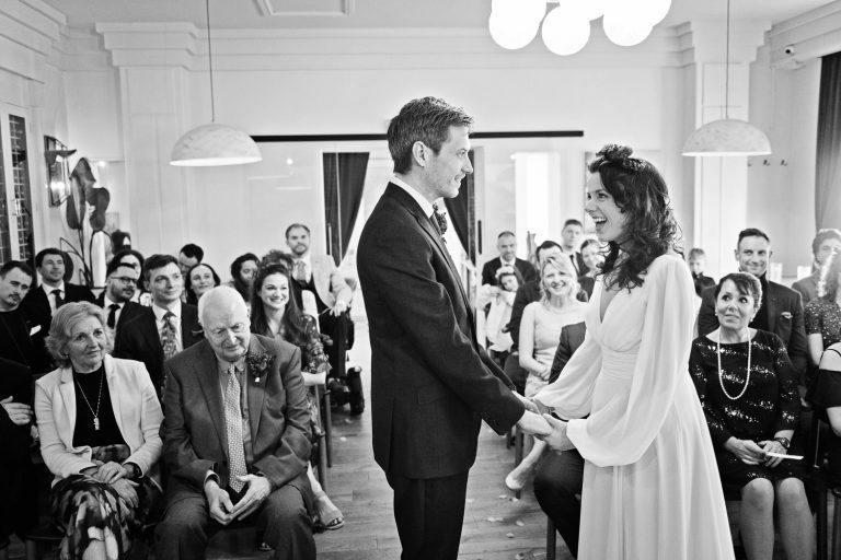 B&W candid photo of bride and groom standing at the front of their wedding ceremony, holding hands and smiling and laughing at each other. Wedding guests are smiling and listening.