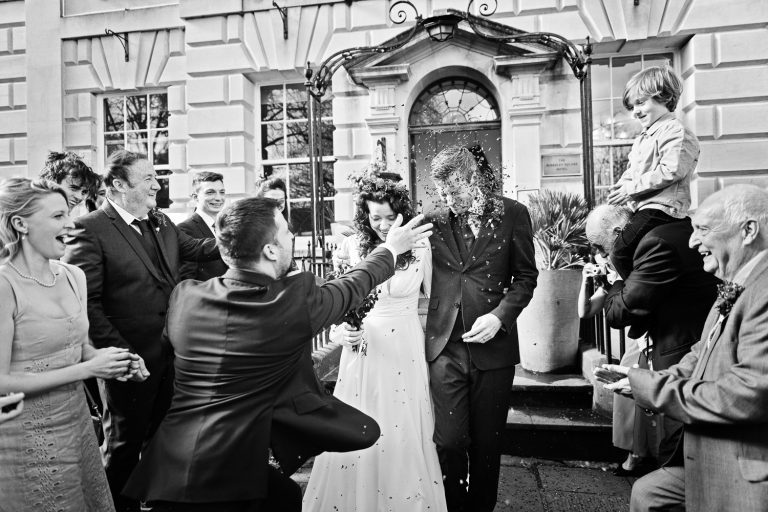 Documentary photo of bride and groom exiting The Berkeley Square Hotel to their close friends and family throwing confetti at them, particularly one guest who is taking aim at the groom with a rather large handful of confetti flowers hitting the grooms face.
