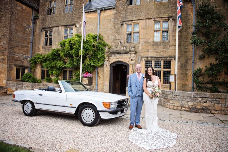 Bride and groom stand in front of the Lygon Arms with the grooms classic car next to him.