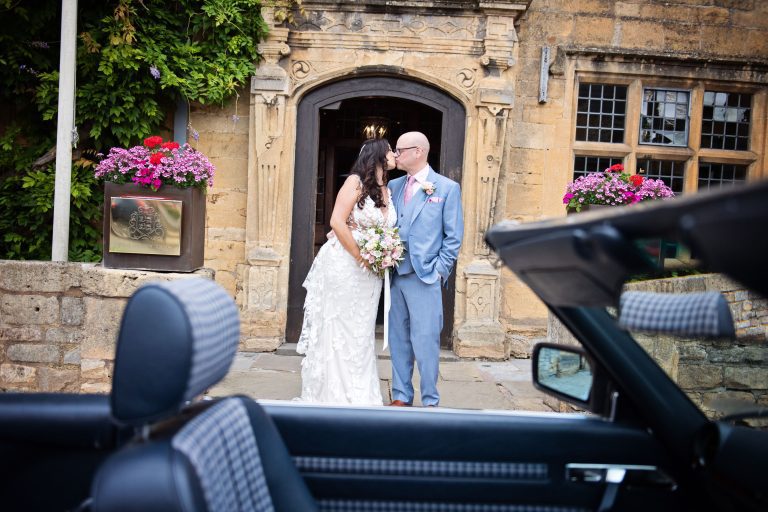 Bride and groom kiss in front of Lygon Arms. The grooms classic car is in front of them.