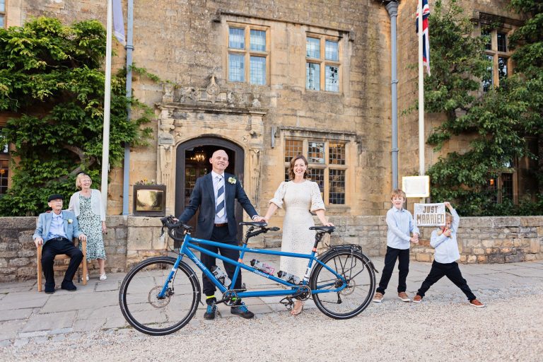Bride and groom stand in front of the Lygon Arms hotel holding their tandem bike, with their pageboys holding a just married sign.