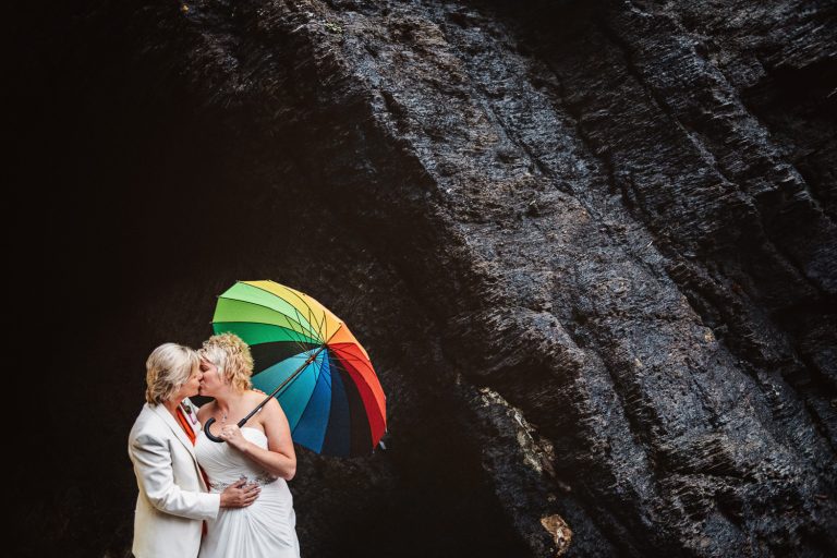Bride and bride kiss with a rainbow coloured umbrella. They are standing next to limestone cliff rocks. Could be an iconic LGBTQ+ image.