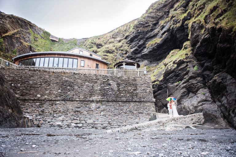 Bride and bride stand with their umbrella in front of Tunnel Beach wedding venue.