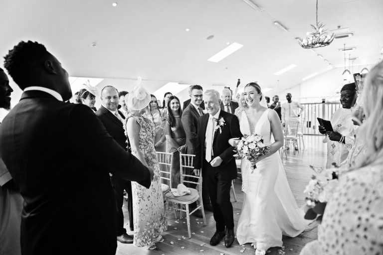 B&W storytelling photo of the bride smiling as she walks down the isle with her father by her side.