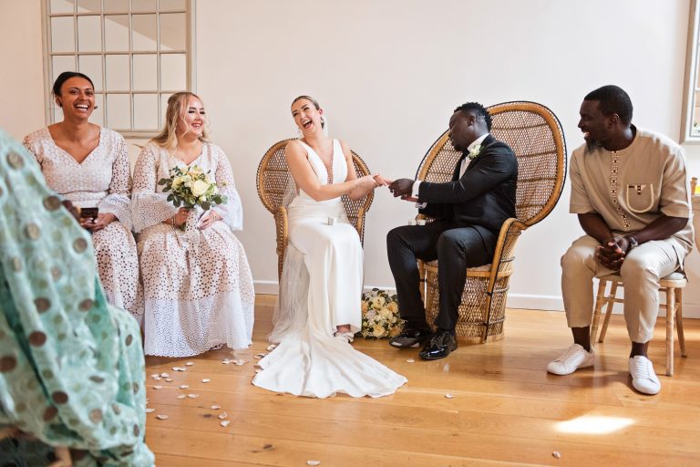 Bride putting the wedding on the grooms finger, watched by close family members. Everyone laughing. Natural.