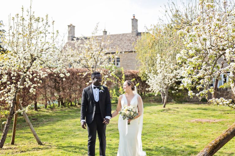 Bride and groom walk hand in hand in front of a blossoming fruit tree.