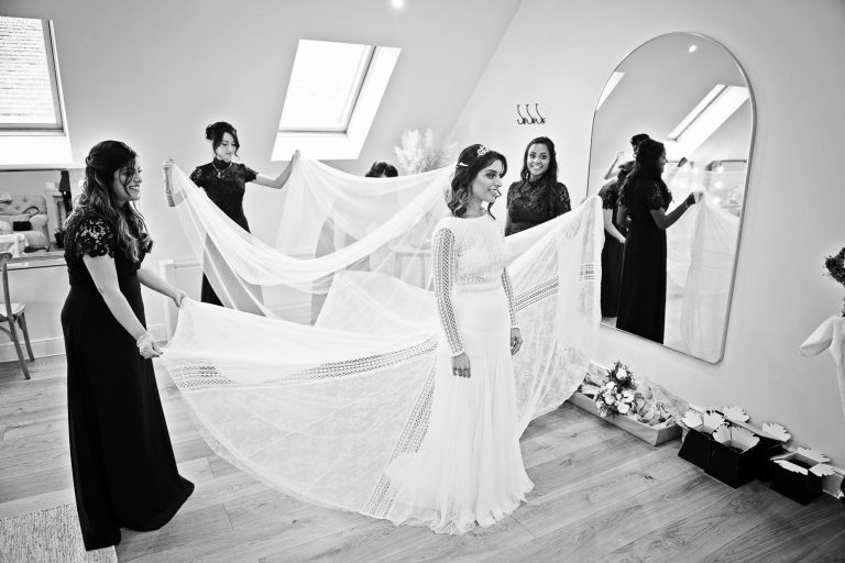 B&W photo of bride in her wedding dress being helped by her bridesmaids.