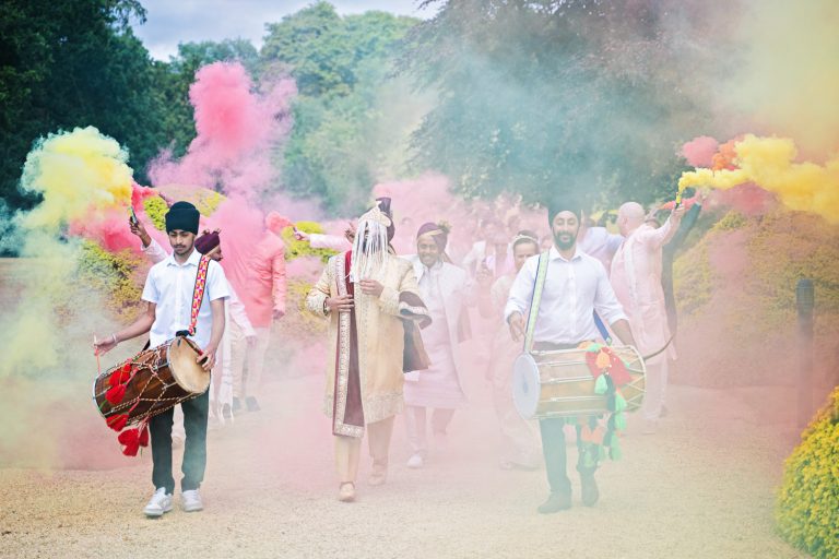 A colourful Baraat, with smokebombs, drums and grooms family members behind, walking towards the entrance of the wedding.
