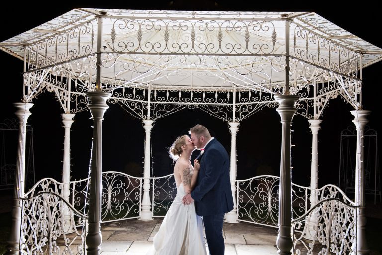 Bride and groom kiss at night underneath the lit bandstand at Manor by The Lake.