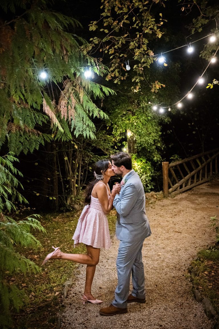 Cute photo of bride and groom kissing at night next to a wooden bridge and underneath festoon lights. Bride's one leg is raised up.