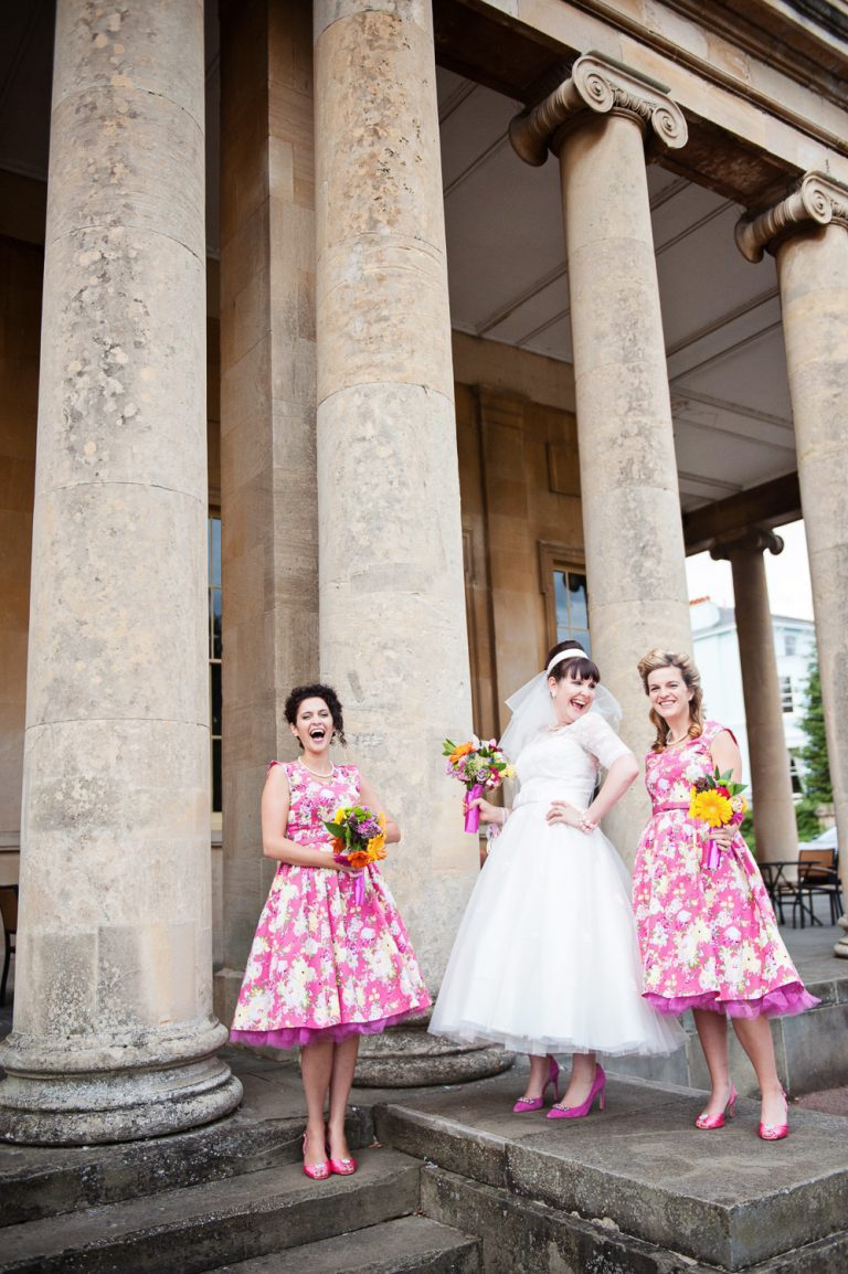 Bride with her bridesmaids in their flowery pic bridesmaids dresses outside the front of Pittville Pump Rooms large pillars.