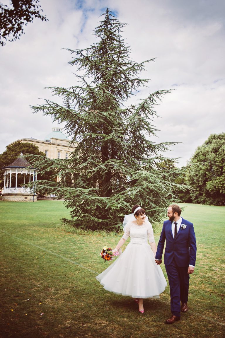 Bride and groom, smile and hold hands as they walk through the Pittville Gardens, part of the Pittville Pump Rooms behind them.