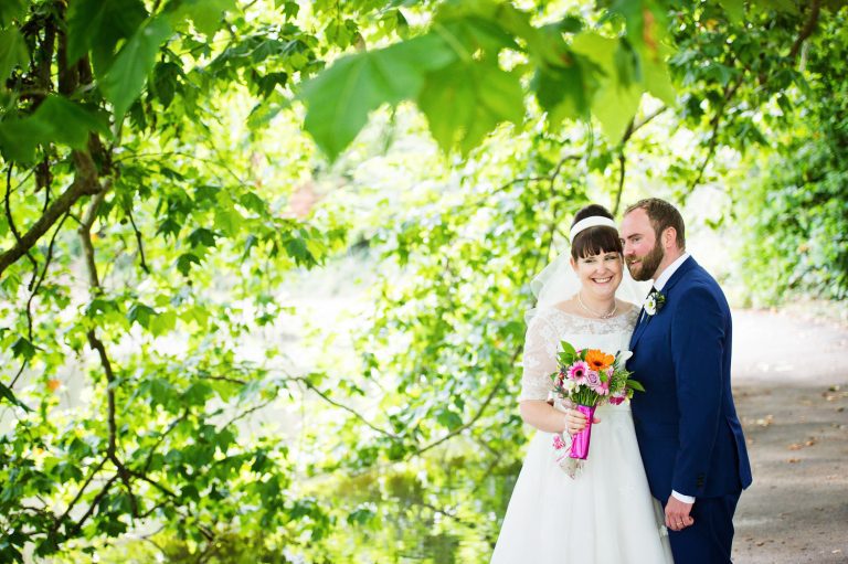 Bride and groom smile under a tree at Pittville Pump Rooms/ Gardens.