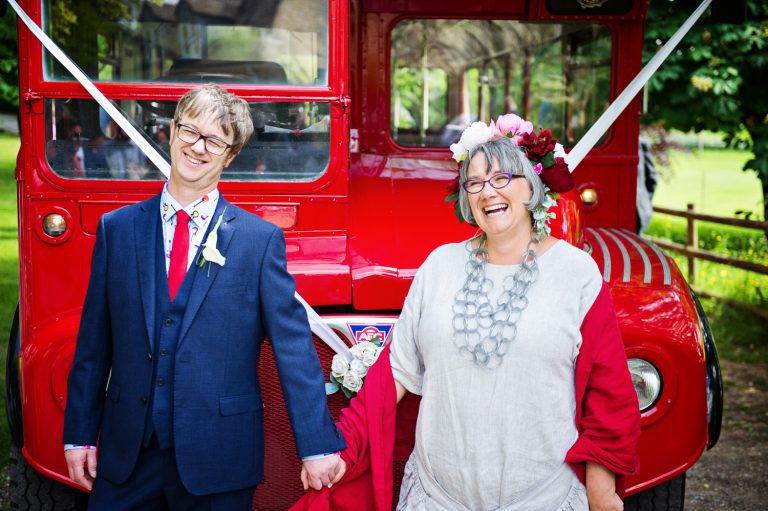 Bride and groom stand in front of a red double decker bus outside Painswick Rococo Gardens.