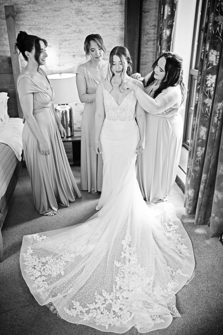 B&W portrait photo of bride having jewelry added by her bridesmaids.