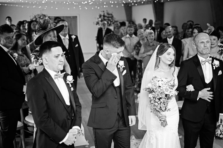 Groom wiping his eye for tears as his bride walks down the wedding isle with the wedding guests behind them.