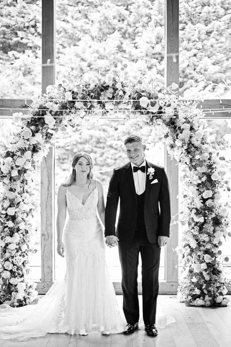 Bride and groom hold hands as they stand in front of a flower arch at The Mill Barns. B&W image.
