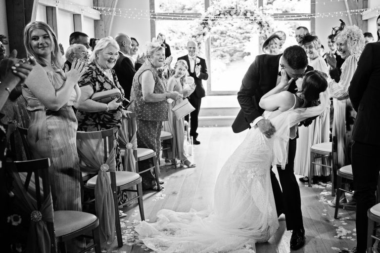 Bride and groom do the dip and kiss in front of wedding guests as they walk down the isle after getting married at The Mill Barns.