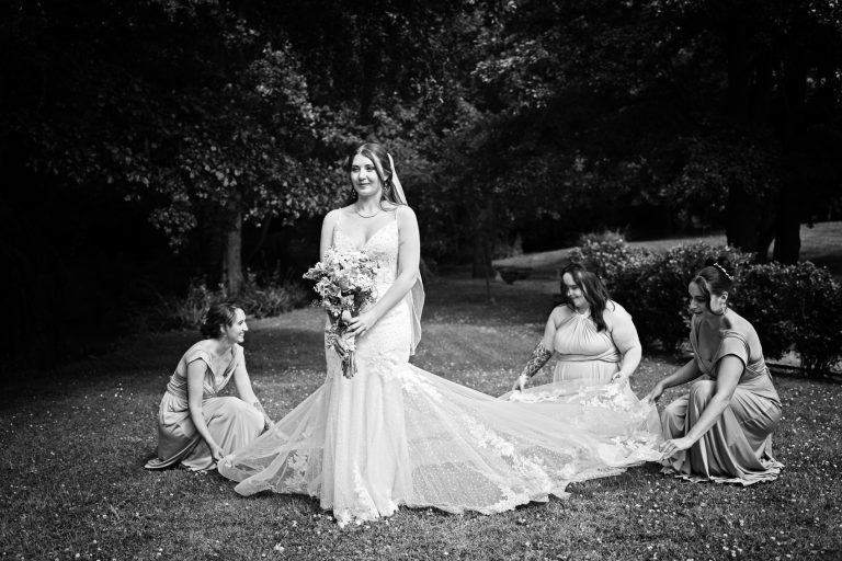 Bride standing, holding her flowers whilst the bridesmaids look after her wedding dress. B&W photo.
