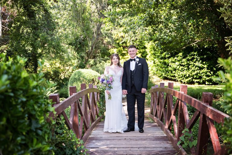 Bride and groom stand side by side on a wooden bridge with green trees and bushes surrounding them.